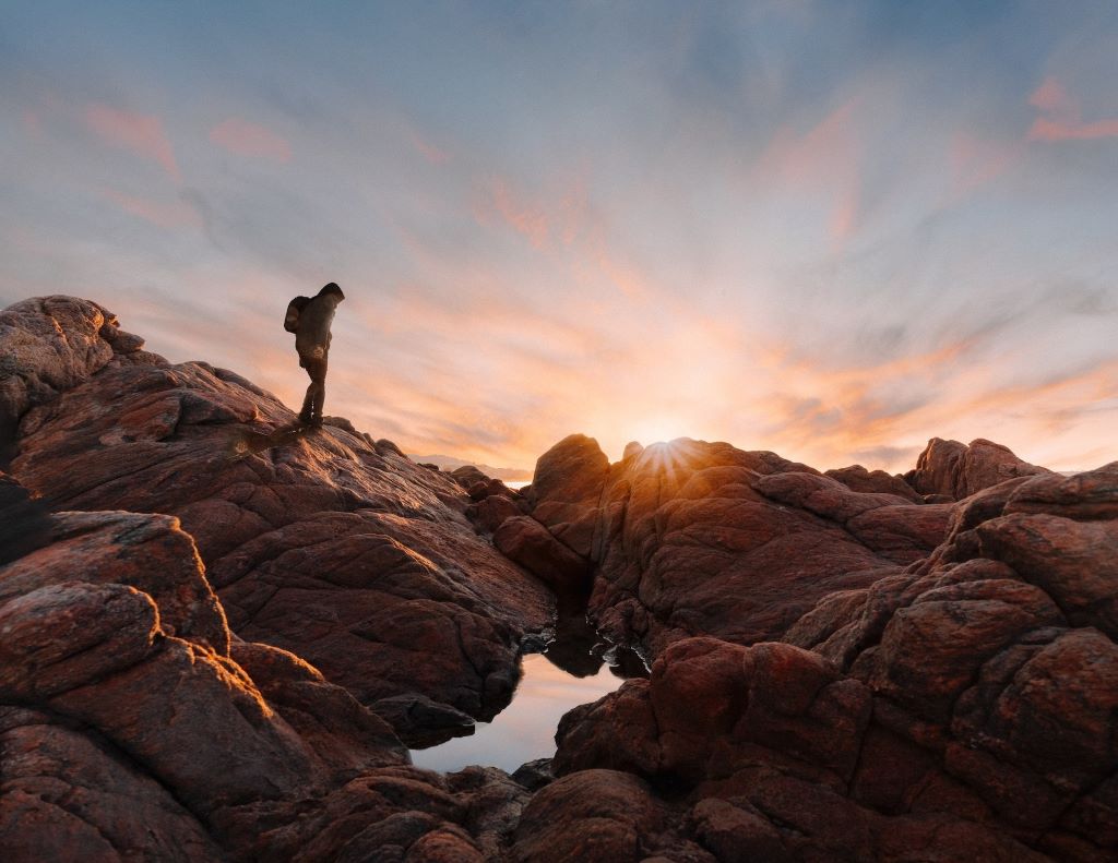A person stands on rocks looking at the sunset. Photo by Samir Belhamra