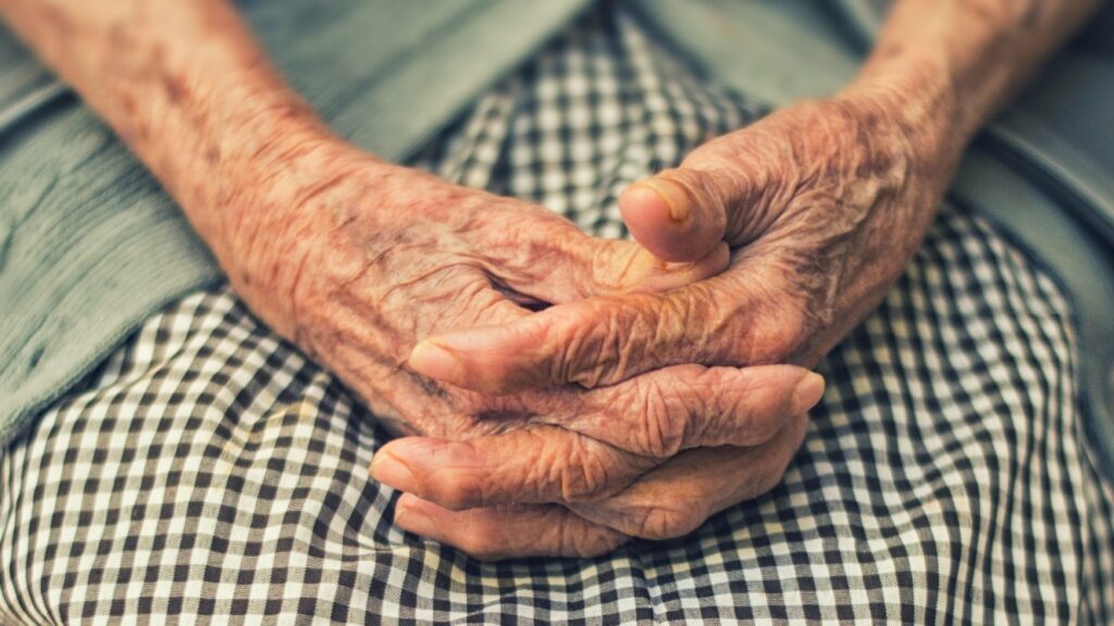 An elderly woman clasps her hands together in her lap