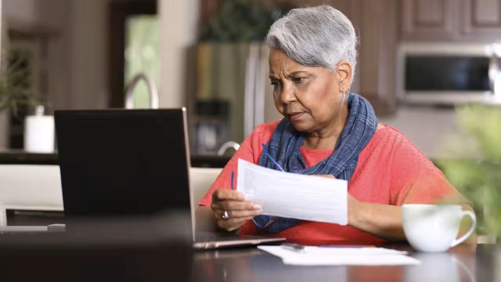 A woman looks worryingly at an open laptop, while holding paperwork